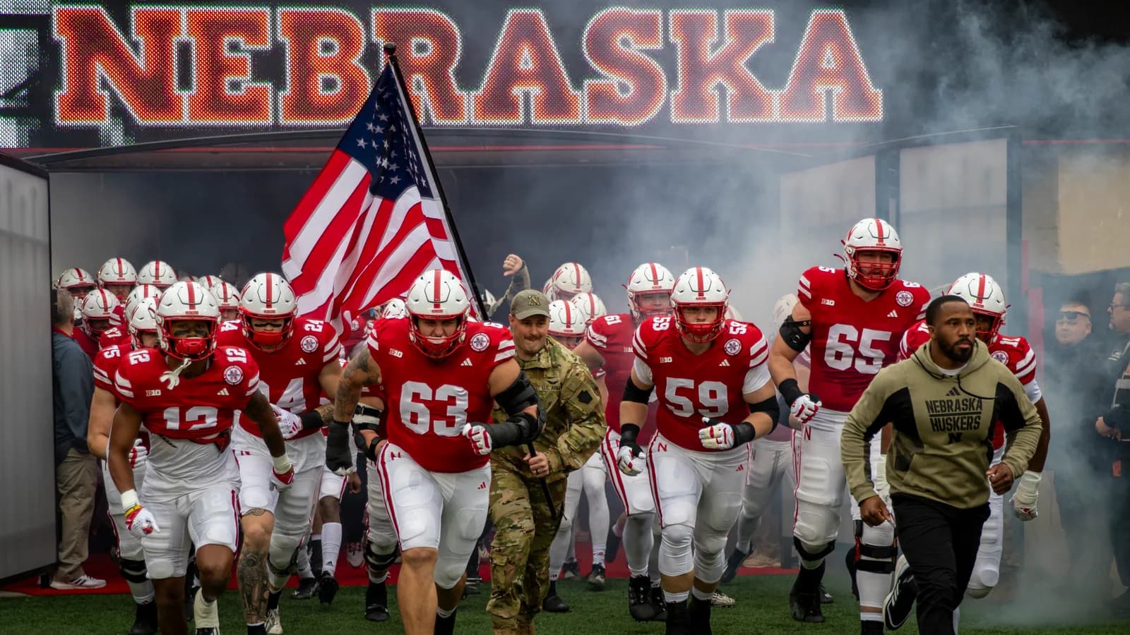 Nebraska football players run onto the field beneath a large Nebraska display.